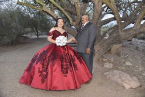 Quinceañera con vestido rojo y detalles negros posando con un hombre en traje gris junto a un árbol grande en un paisaje desértico, foto de AZ Foto y Video / IAZ Photo Studio.