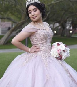 Quinceañera con vestido rosa claro y tiara posando con ramo de flores en un parque.