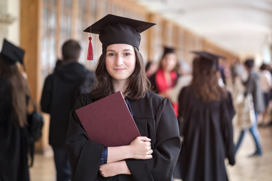 Graduada con toga y birrete sosteniendo una carpeta frente a un pasillo universitario con otras personas al fondo.