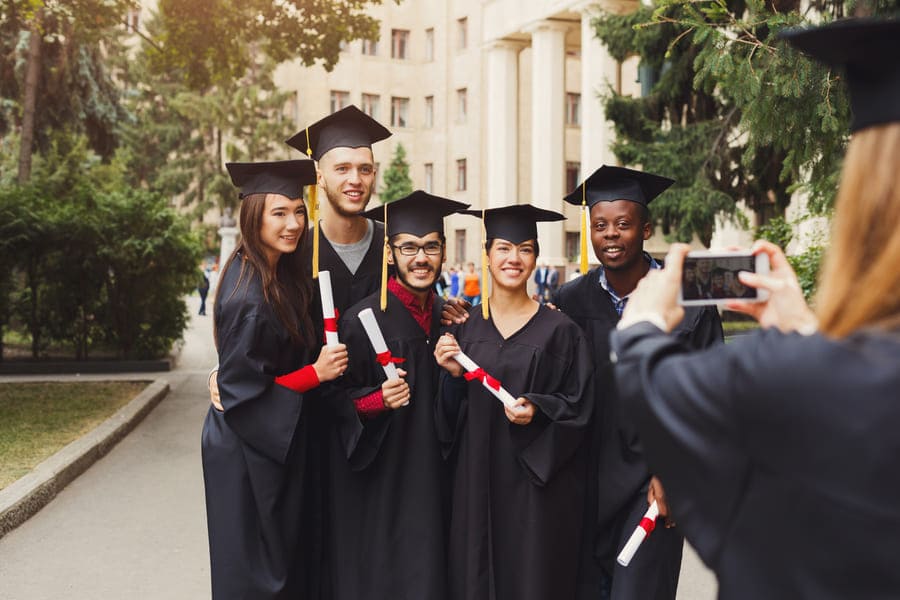 Grupo de graduados con toga y birrete posando con diplomas en un campus universitario mientras otra persona les toma una foto con el celular.