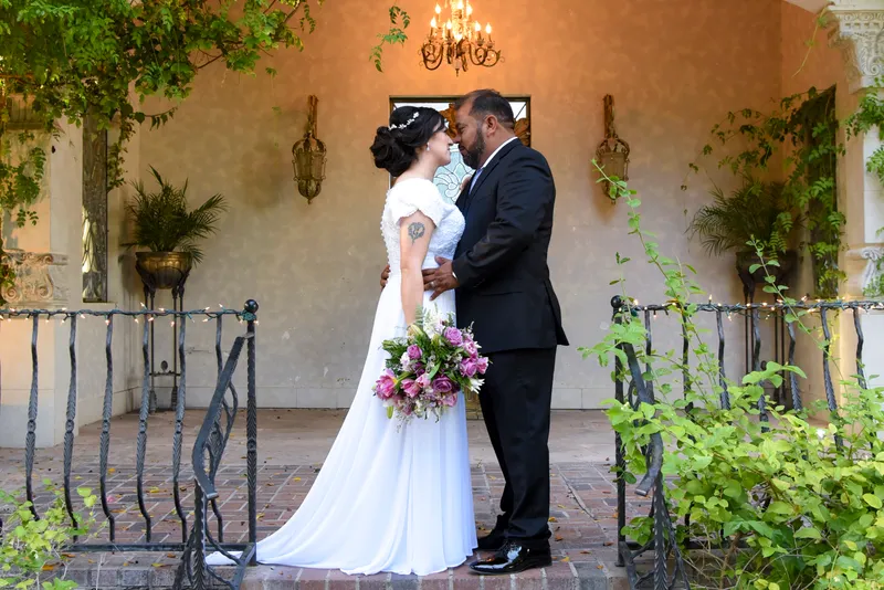 Novia con vestido blanco y novio con traje oscuro abrazados en la entrada de una elegante residencia.