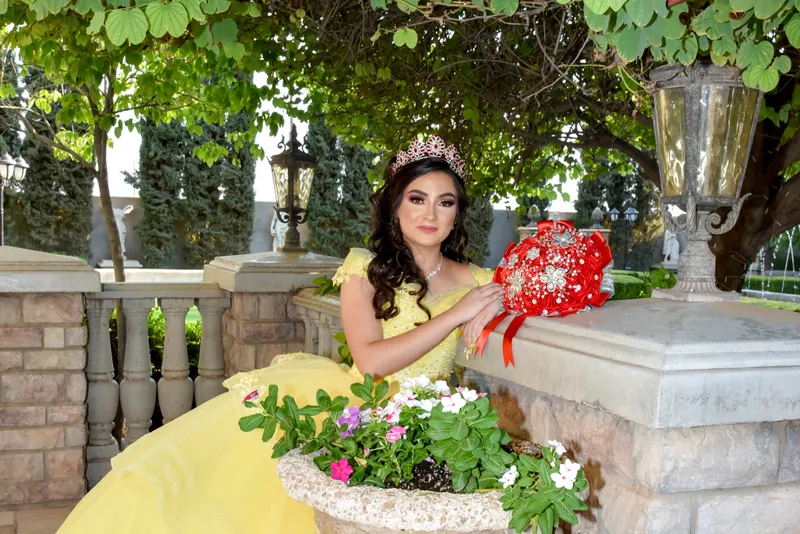 Quinceañera con vestido amarillo brillante, corona y ramo de flores rojas posando en un jardín en Gilbert AZ.