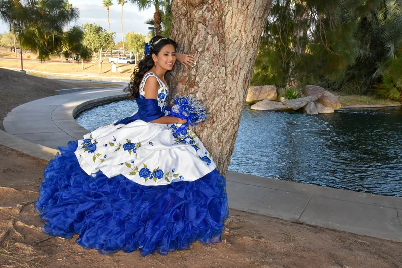 Quinceañera con vestido azul y blanco posando junto a un árbol grande frente a un lago en Gilbert.