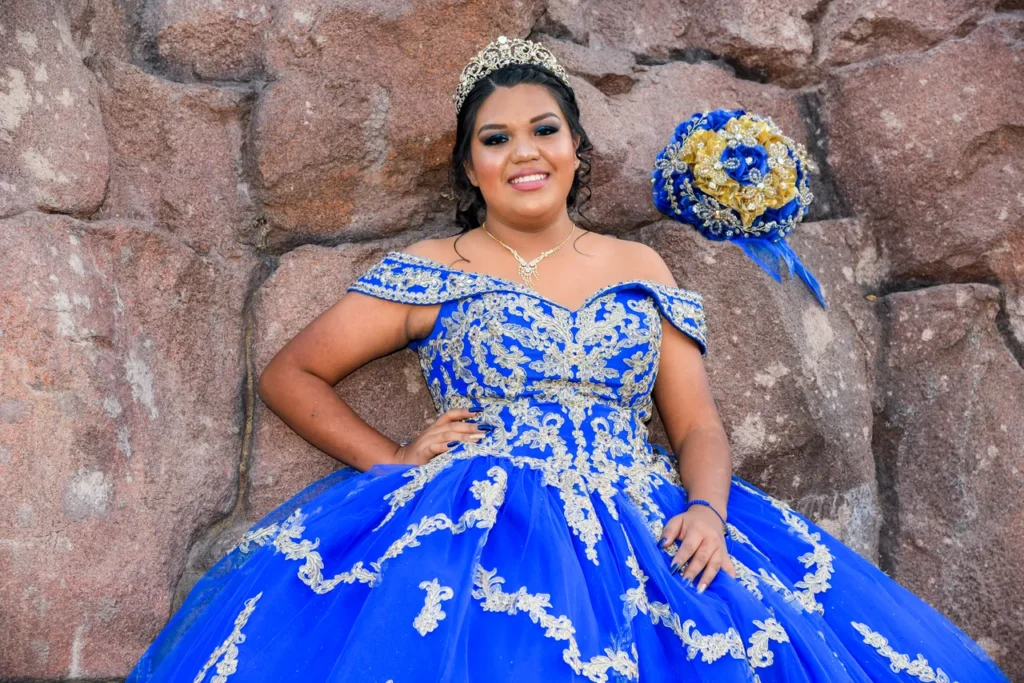Quinceañera posando con vestido azul real y ramo de flores frente a una pared de piedra.