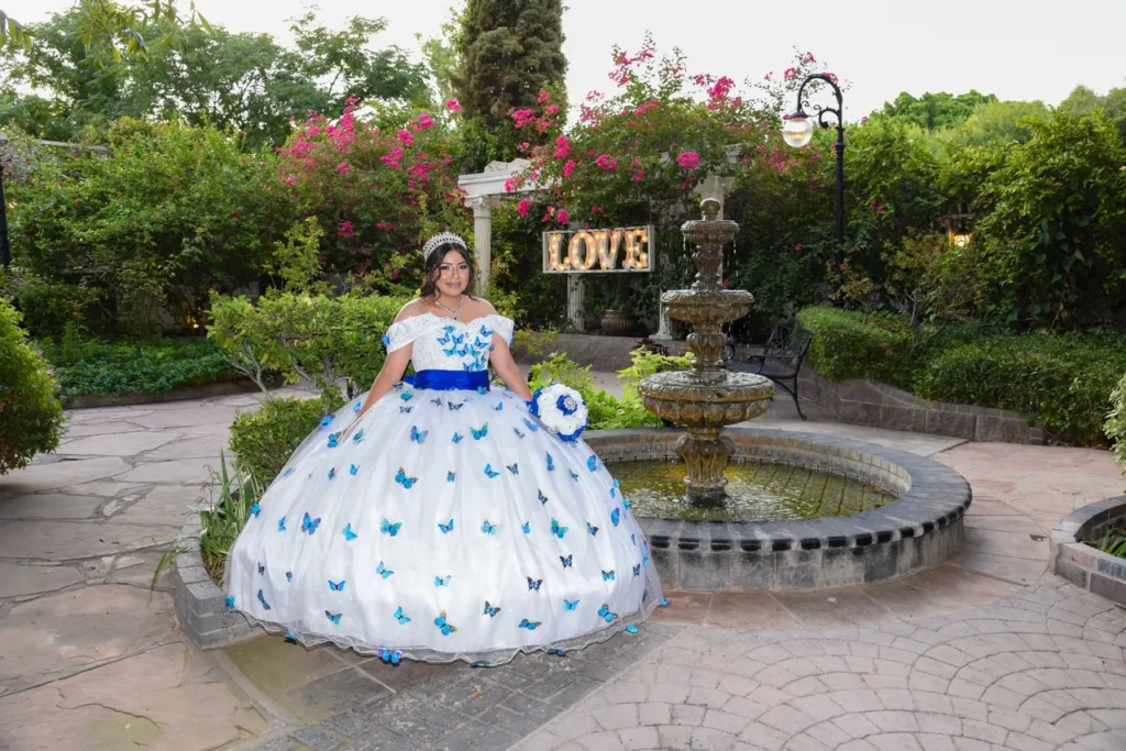 Quinceañera con vestido blanco y mariposas azules posando frente a una fuente de piedra en jardín.