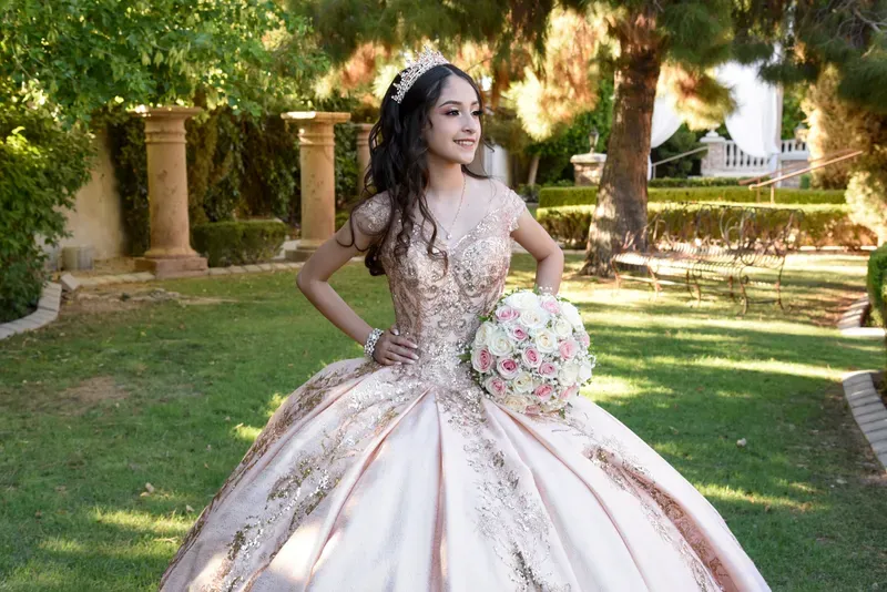 Quinceañera con vestido rosa brillante y ramo de flores posando en un jardín iluminado por el sol.